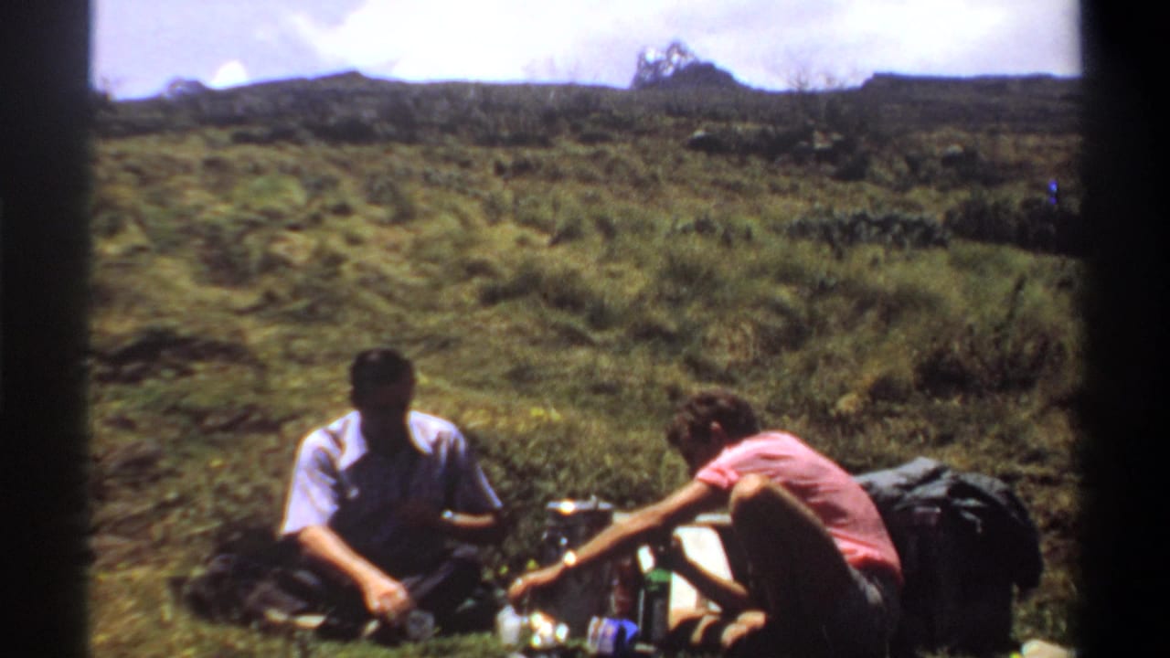 Two men sit on a grassy hillside with bottles and packs nearby.
