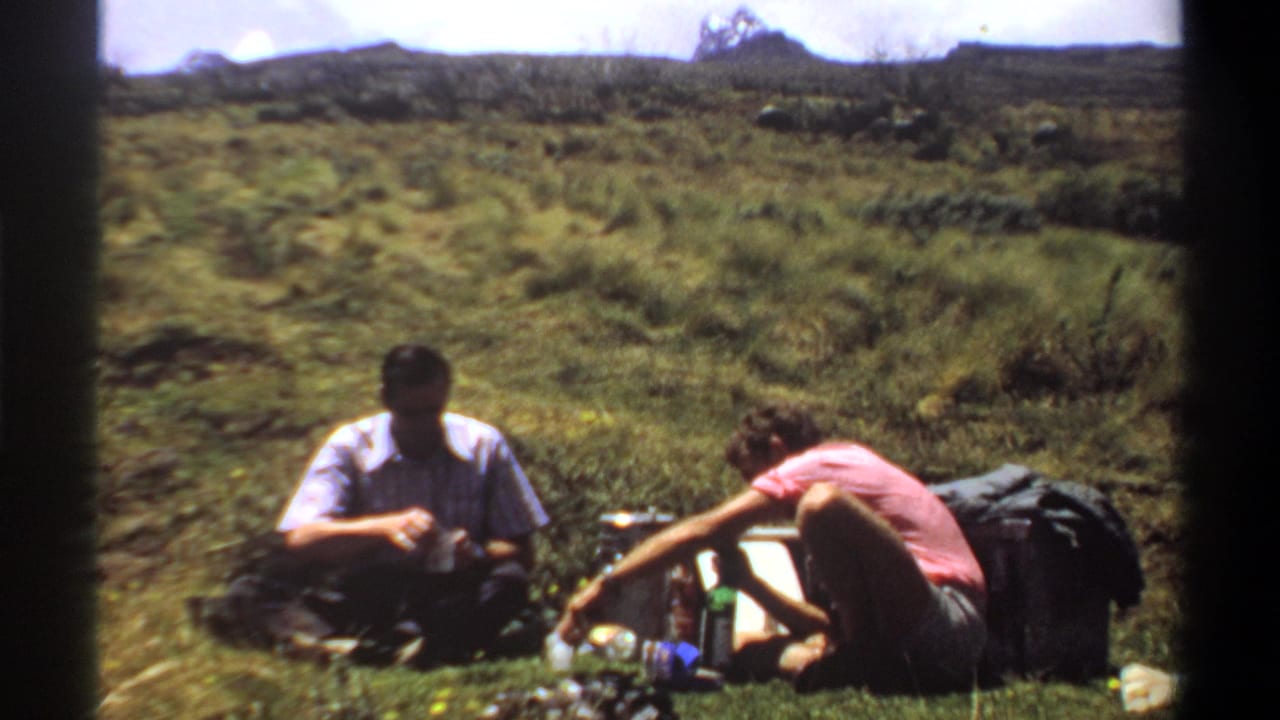 1969 Khartoum-area home-movie frame of two adult men seated outdoors, surrounded by packs and bottles on a sunlit grassy slope.