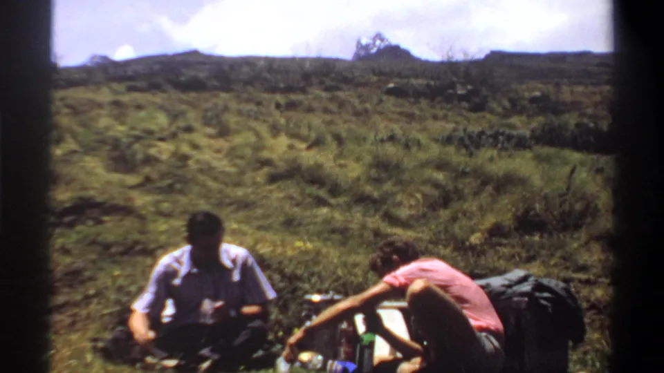 Faded 8mm frame from 1969 Sudan showing two men seated near supplies on a grassy slope, with open terrain and sky behind them.