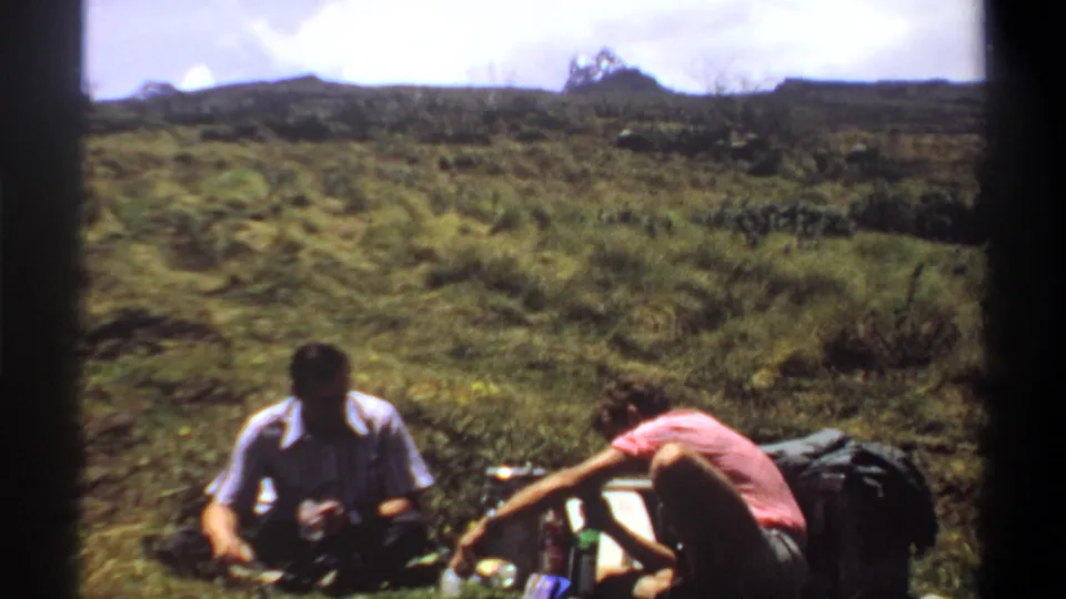1969 Khartoum home-movie frame of two adult men relaxing outdoors, with drink containers and bags arranged beside them on the grass.