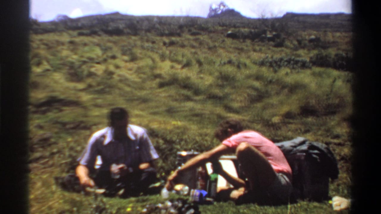 In 1969 Khartoum-area home-movie footage, two adults sit on grass with picnic supplies in an open field.