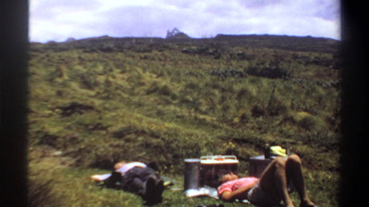 1969 8mm home-movie frame from Khartoum, Sudan, showing two adults resting in green pasture near picnic items and a distant hilltop structure.