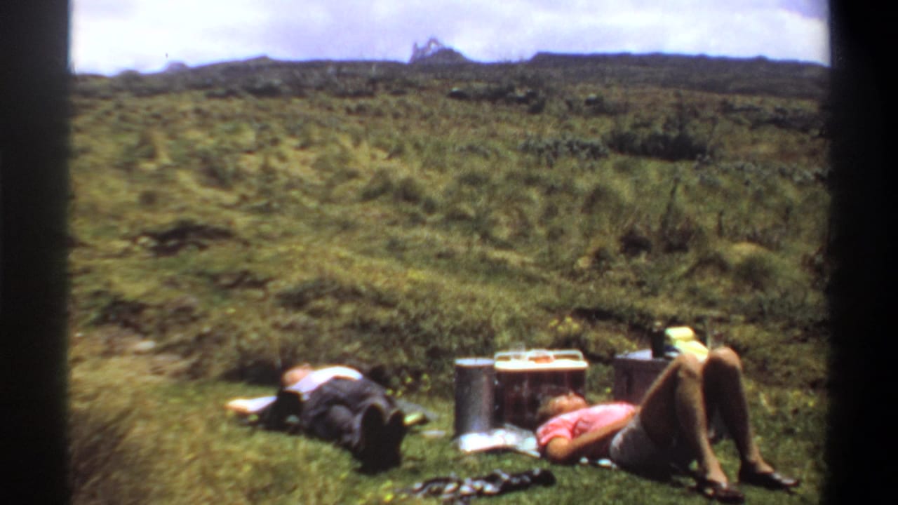 1969 Khartoum home-movie frame showing two adults relaxing on grass beside a cooler in an open green landscape.