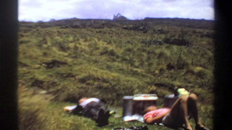 1969 Khartoum frame with two adults reclining on a grassy hillside beside a cooler and thermos, filmed in soft 8mm color.