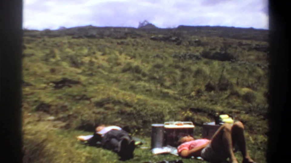 Two adults rest low in the frame during a 1969 Khartoum home-movie shot, with picnic objects and a broad field stretching behind them.