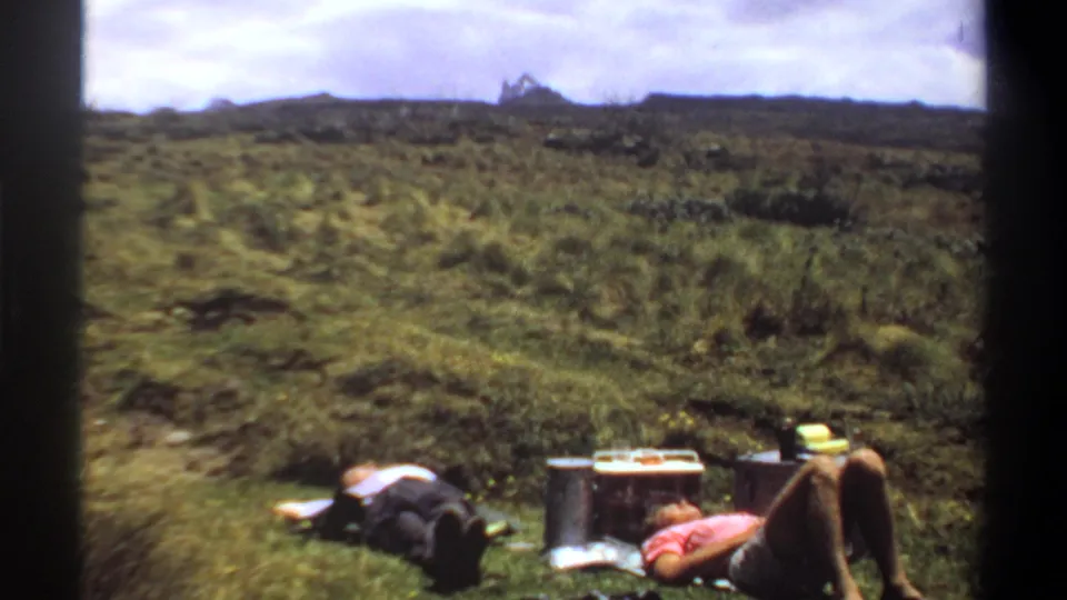 1969 Khartoum home-movie still showing two adults relaxing beside picnic items in a green field beneath a cloudy sky.