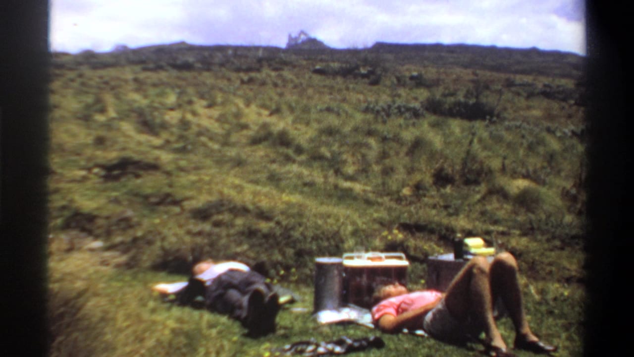 1969 Khartoum home-movie frame showing two people resting on grass near supplies, with scrubby hillside and distant ridge behind them.