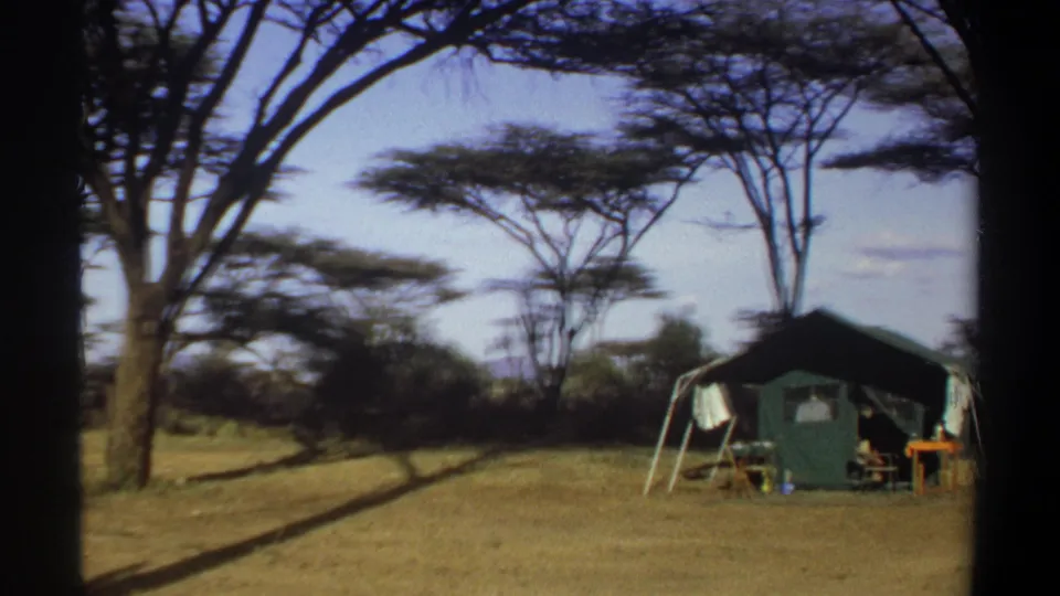 1969 Khartoum home-movie frame showing a green canvas camp tent under tall trees, with laundry and camp furniture outside.