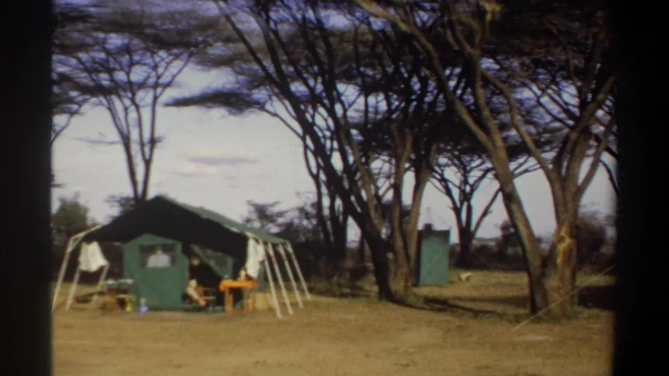 1969 Khartoum frame showing the tent camp at left, a small blue-green structure among trees, dry grass, and camp furnishings.