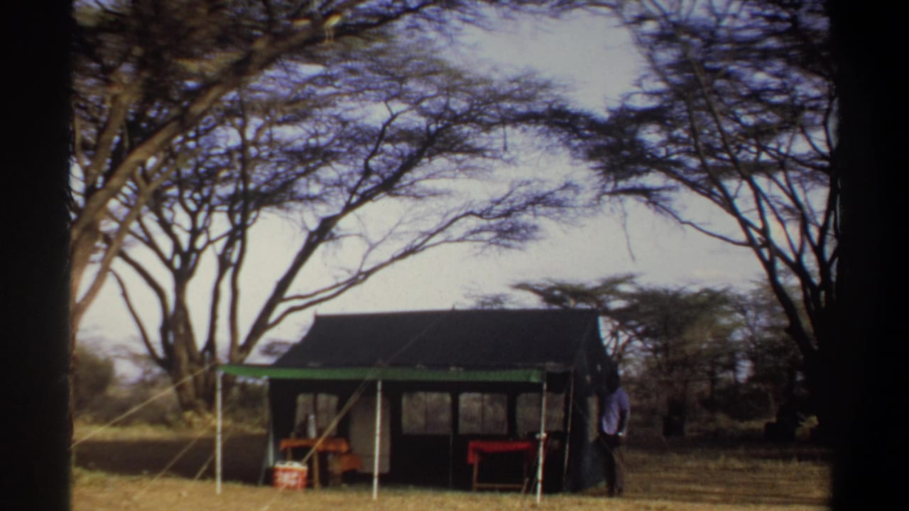 1969 Sudan travel footage frame with a man beside a canvas tent, camp tables, and trees in the background.