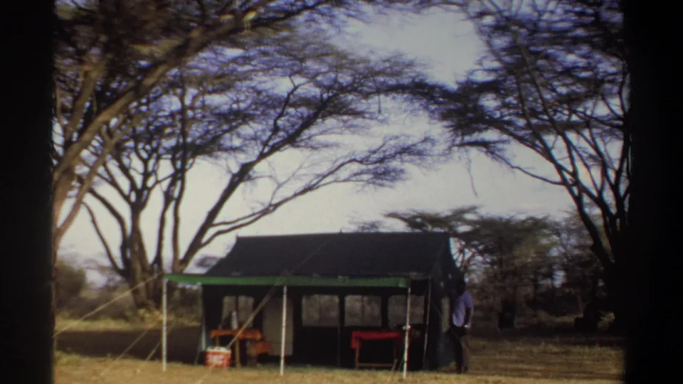 1969 Sudan travel footage frame with a man beside a canvas tent, camp tables, and trees in the background.