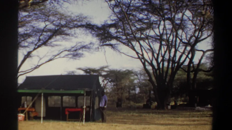 The 1969 Sudan camp pan shows the tent edge, a standing man, dry grass, and a broad canopy of trees.