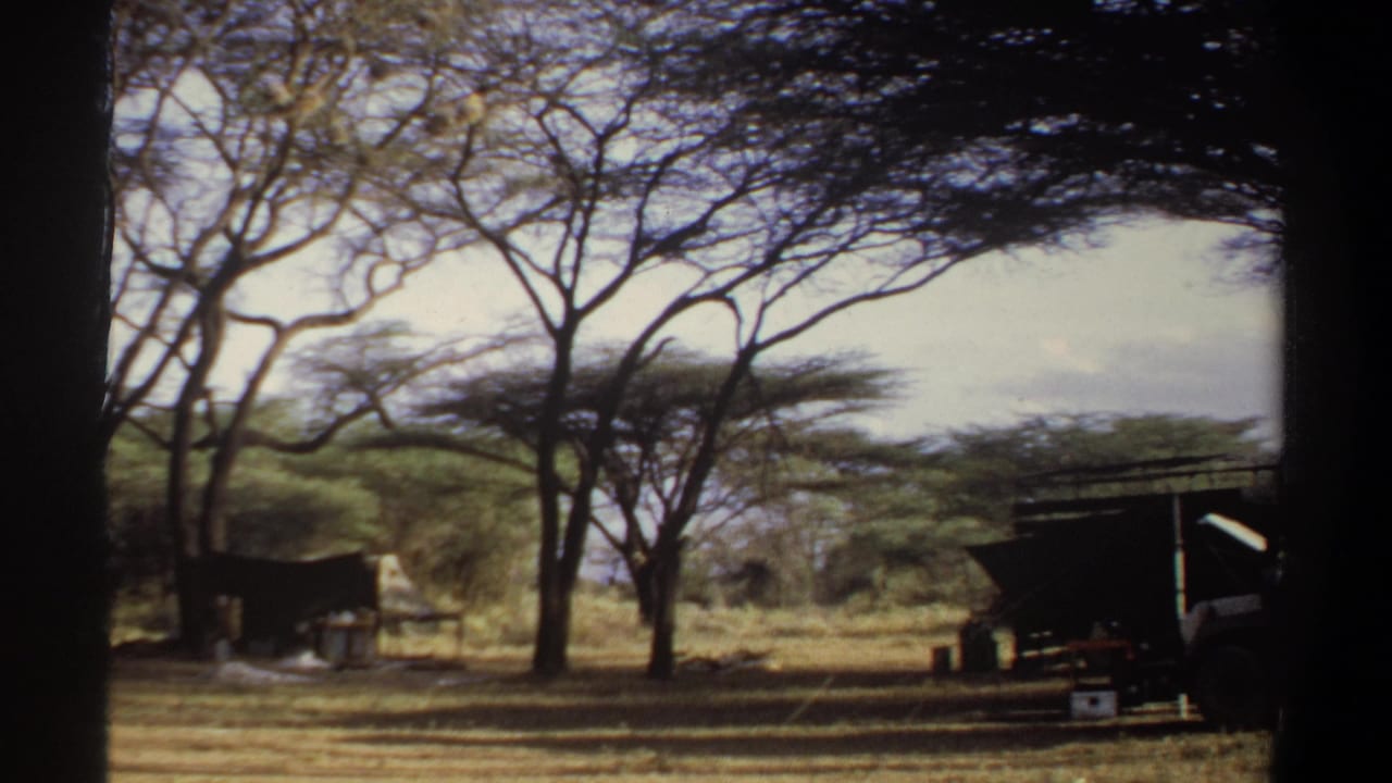 1969 Khartoum frame with acacia-like trees, dry ground, a shelter, and the edge of a vehicle at the right.