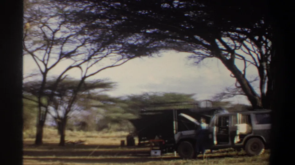 1969 Khartoum home-movie frame showing an open safari-style vehicle, raised hood, shelter, boxes, and a standing adult.