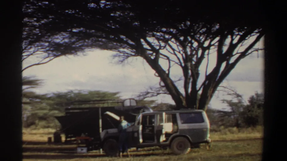 1969 Khartoum frame showing a boxy vehicle with open doors under a large tree, with an adult near the camp setup.