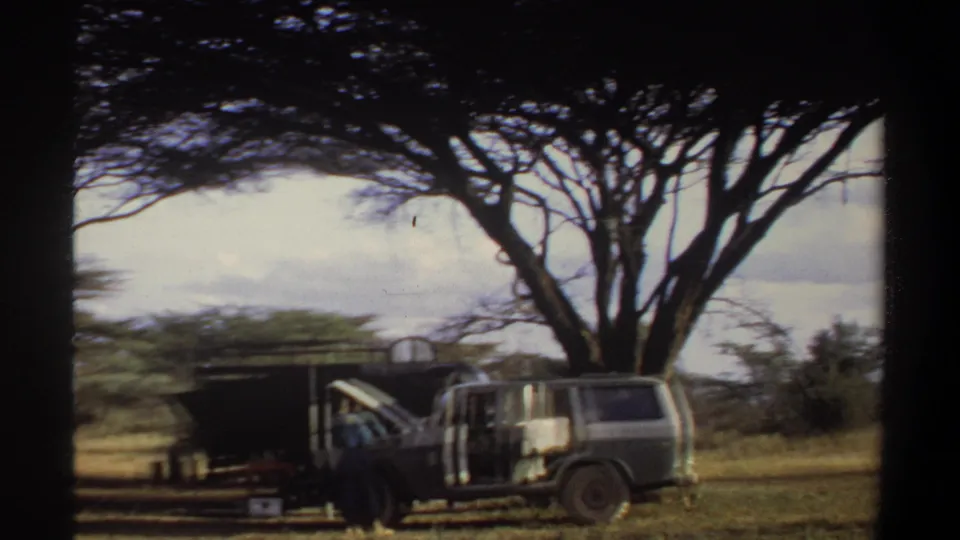 1969 Khartoum-area 8mm frame showing a parked vehicle and dark outdoor shed shaded by a large acacia tree.
