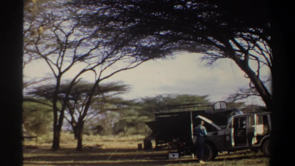 1969 Khartoum-area frame showing a distant person beside a parked vehicle and shaded outdoor shed under acacia trees.