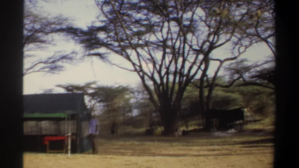 Khartoum-area safari camp frame showing a tent-like structure with green trim, nearby furniture, ropes, and trees in 8mm footage.