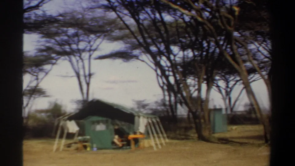 1969 Khartoum, Sudan frame showing a green canvas tent, simple camp furnishings and tall trees across a dry campsite.