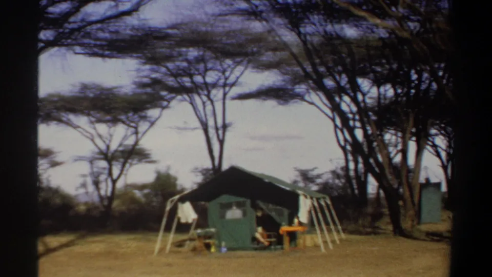 1969 Sudan home-movie frame showing a green safari-style tent under acacia-like trees, with chairs and a small table on dry grass.