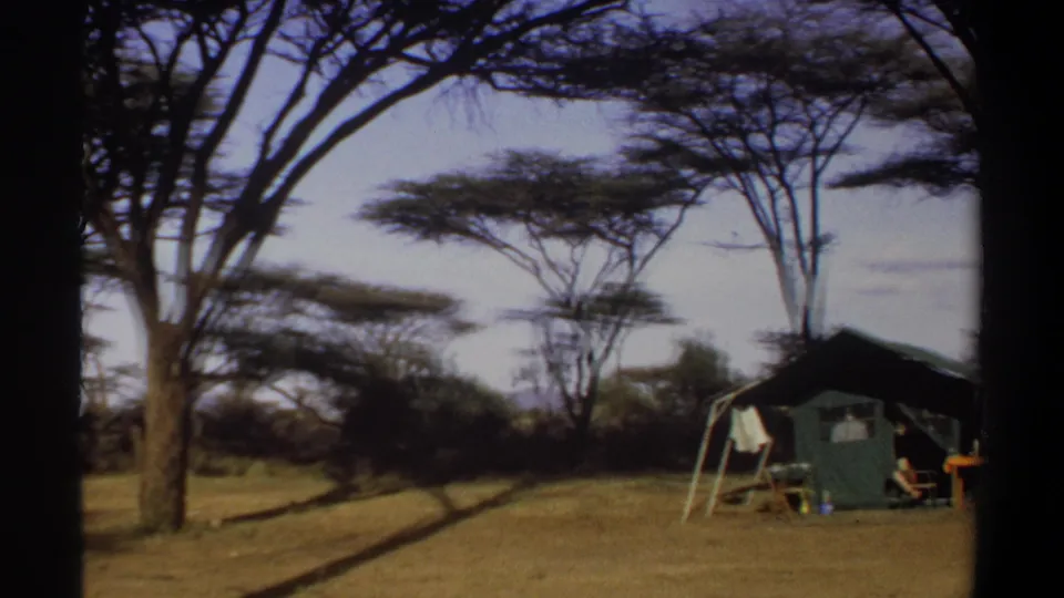 1969 Sudan frame with a green camp tent at the edge of a dry clearing, surrounded by acacia-like trees and faint seated figures.