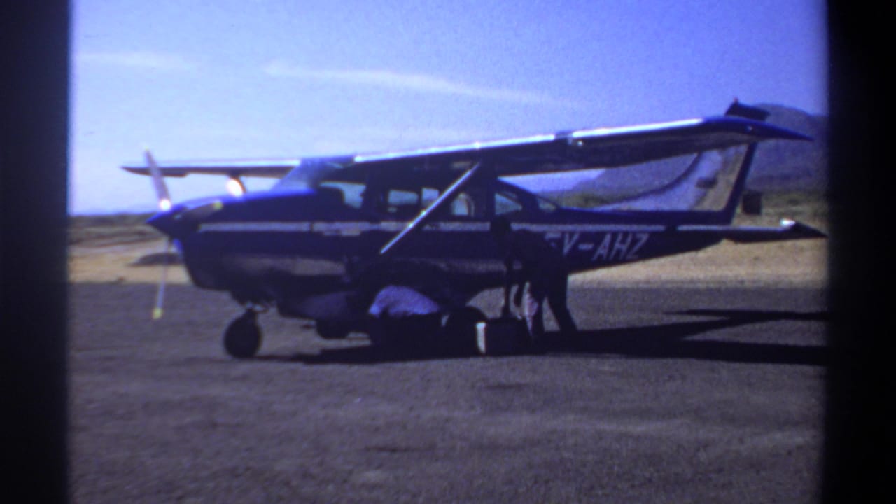 Small blue and white aircraft on a Khartoum airfield in 1969, with one person crouched near the landing gear under a bright sky.