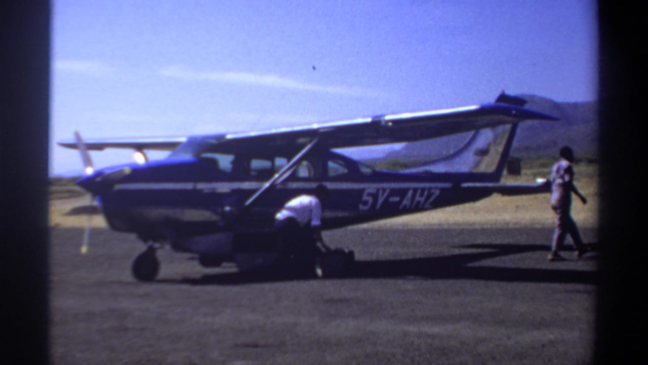 1969 Khartoum home-movie frame showing a blue-and-white single-engine airplane on a dusty airfield with men near the nose and cabin.