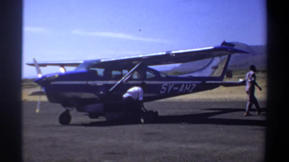 1969 Khartoum home-movie frame showing a blue-and-white single-engine airplane on a dusty airfield with men near the nose and cabin.