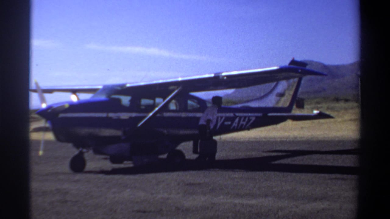 1969 Khartoum frame showing a blue aircraft marked 5Y-AHZ with a person standing near the side door on an open airfield.