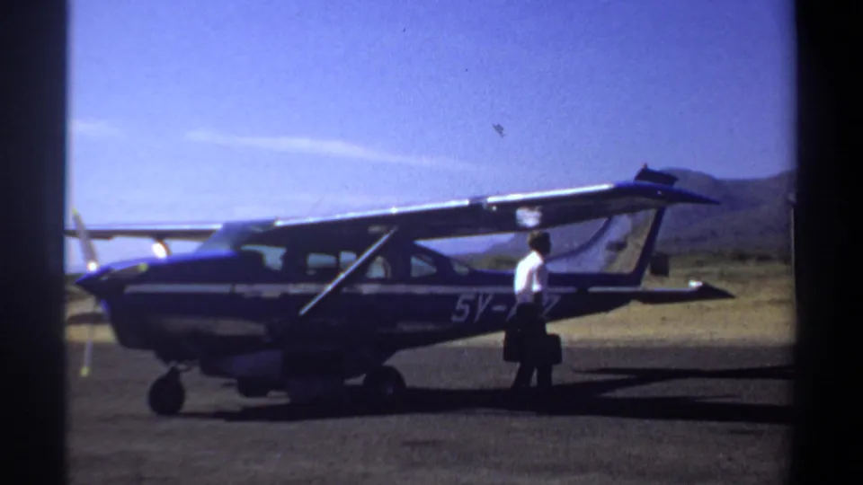 1969 Khartoum home-movie frame with a man in a white shirt carrying a dark bag beside a parked blue single-engine airplane.