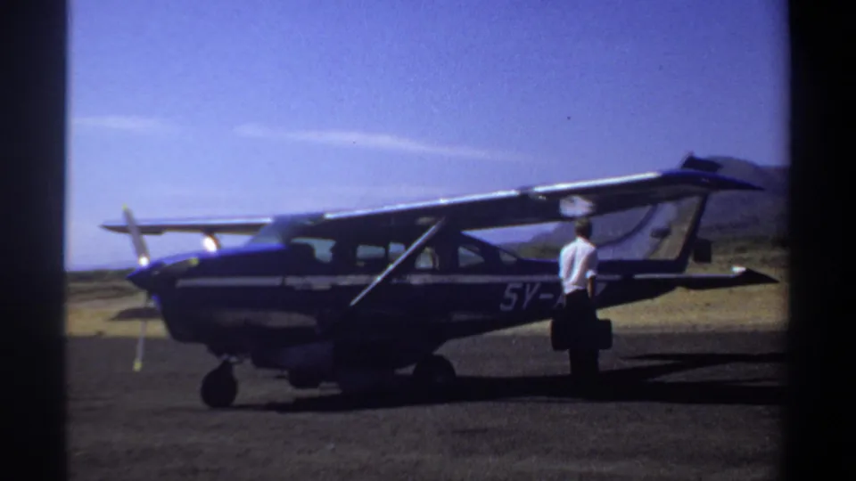 1969 Khartoum frame showing a man standing beside the rear fuselage of a blue-and-white aircraft under a bright sky.