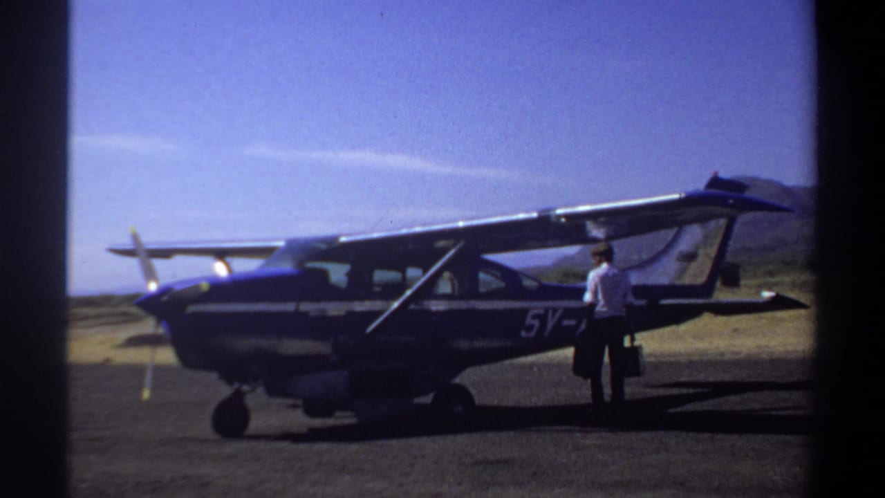 1969 Khartoum home-movie frame showing a parked blue-and-white single-engine airplane and an adult man with a suitcase near the wing.
