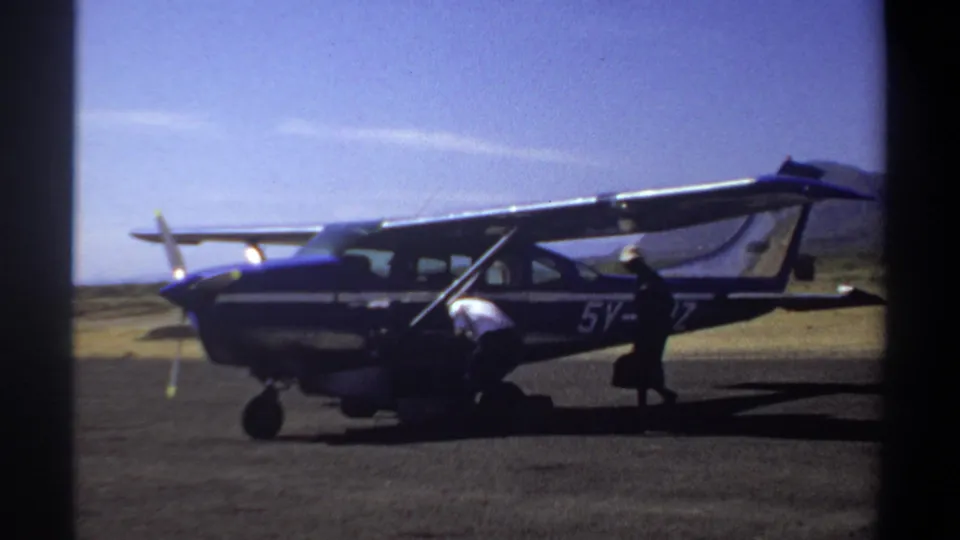 1969 Khartoum home-movie frame showing two adults beside a small aircraft as one bends under the wing near the landing gear.