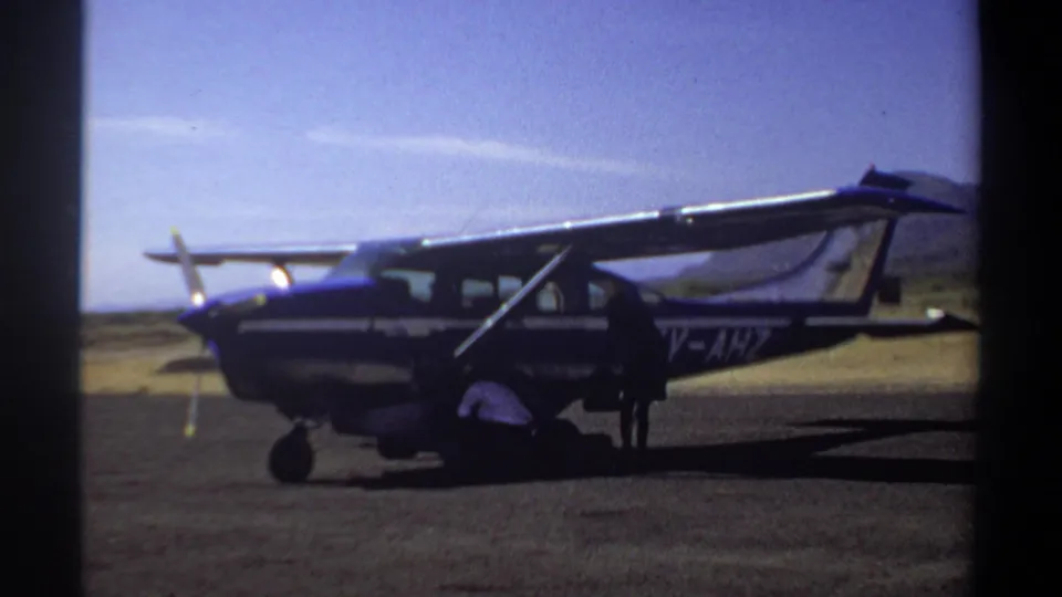 1969 Khartoum frame showing a person ducking beneath the wing of a parked single-engine aircraft on a bright runway.