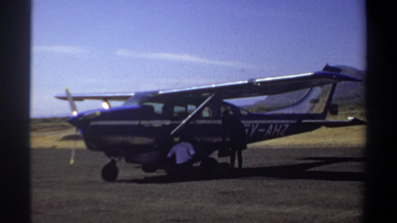 1969 Khartoum home-movie frame showing a small blue and white propeller aircraft on an airfield, with men working near the landing gear and door.
