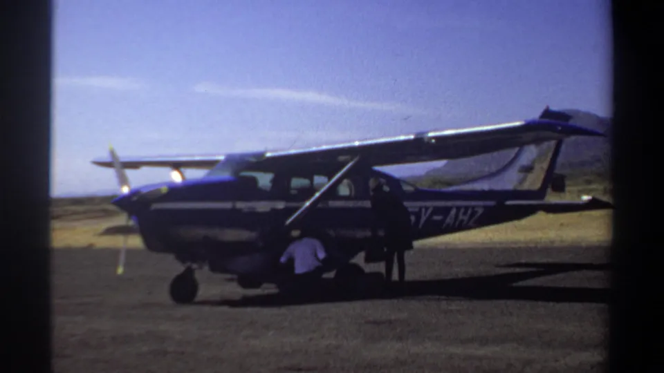 1969 Khartoum home-movie frame showing a small blue and white propeller aircraft on an airfield, with men working near the landing gear and door.
