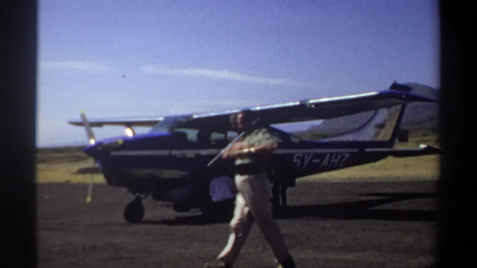 A 1969 Sudan home-movie frame shows a man crossing the foreground in front of a small parked aircraft as workers tend to the plane.