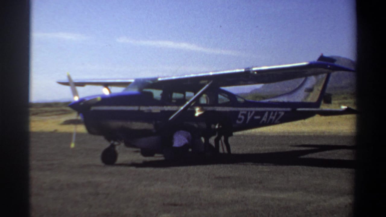 1969 Khartoum home-movie frame showing a small aircraft marked 5V-AHZ with a person crouched beside the landing gear.
