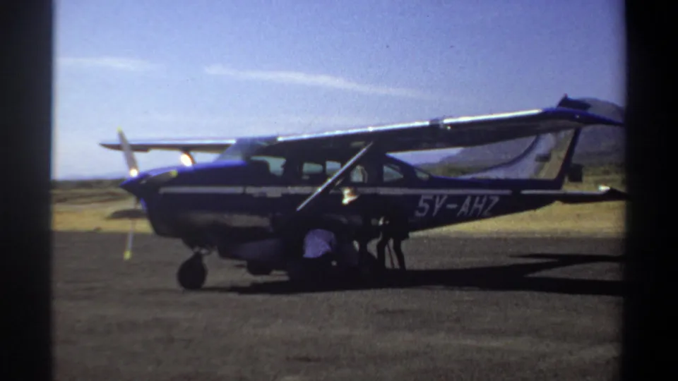 1969 Khartoum home-movie frame showing a small aircraft marked 5V-AHZ with a person crouched beside the landing gear.