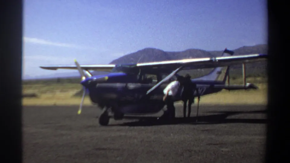 1969 Sudan home-movie frame showing a person beside a small aircraft, with dry airfield terrain and distant hills behind it.