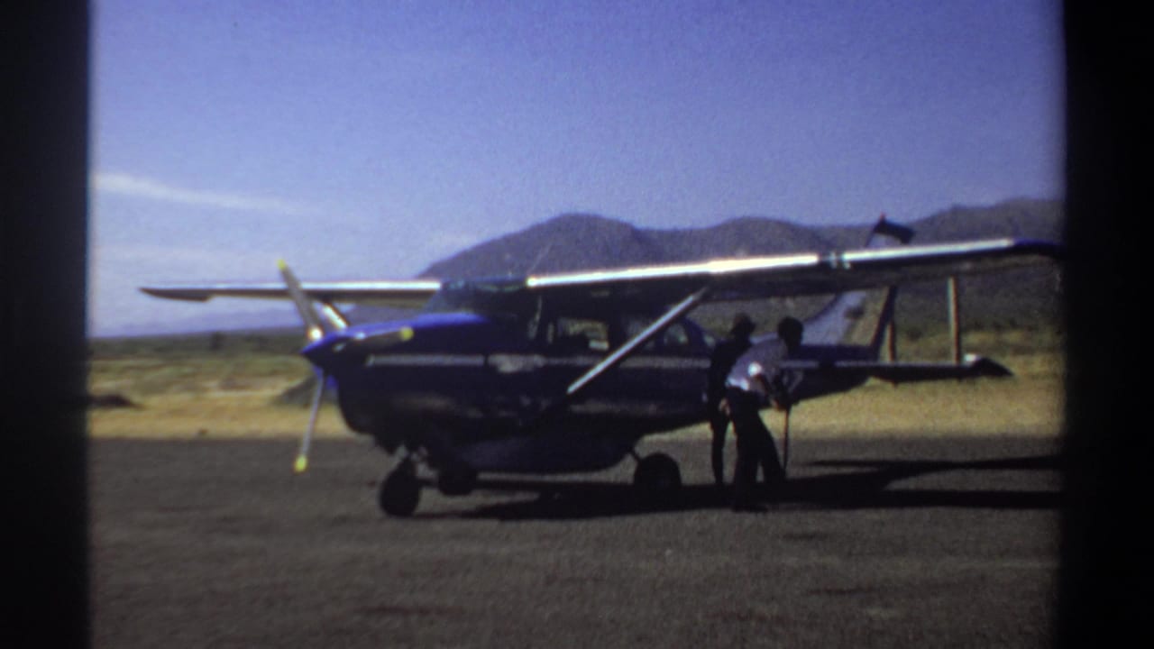 1969 Khartoum home-movie frame showing a blue and silver small propeller plane on an arid airfield as two men stand near the tail section.