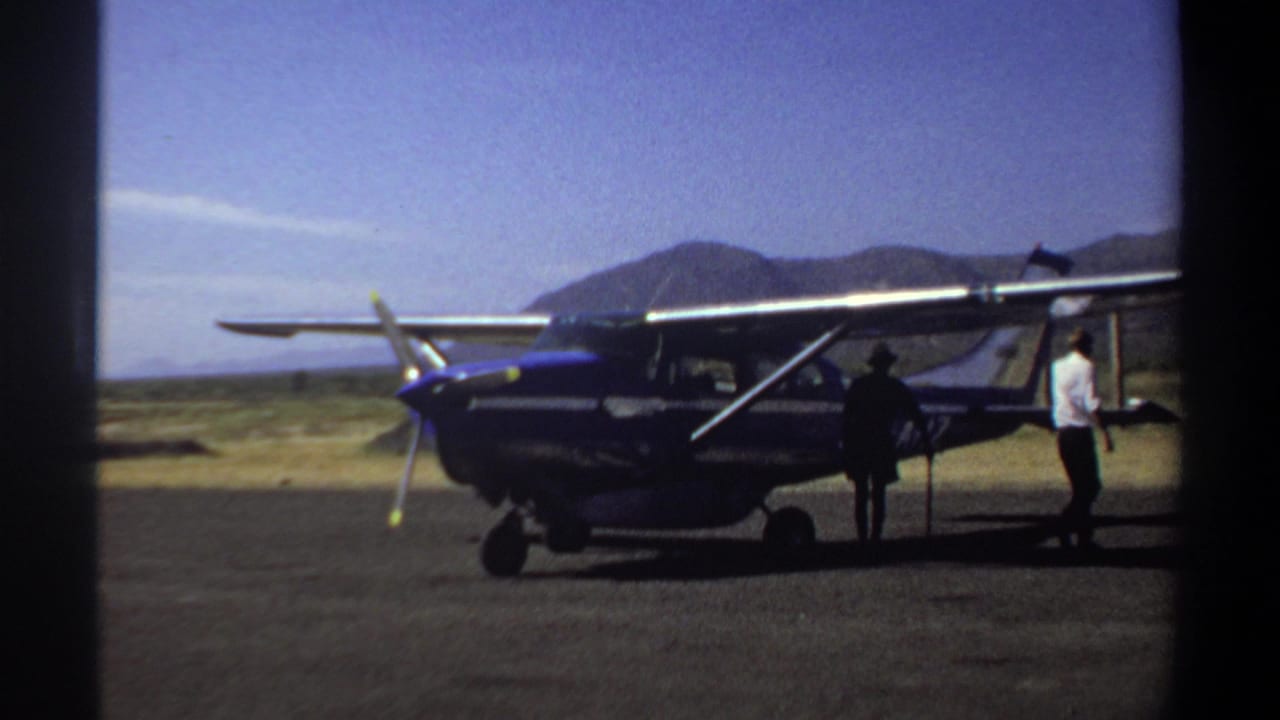 Two men stand beside the rear of a blue propeller plane on an open airstrip.