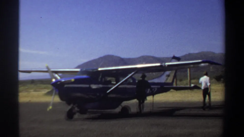 1969 Khartoum 8mm frame showing a white-shirted man near the aircraft rear and a second figure beside the blue high-wing plane.