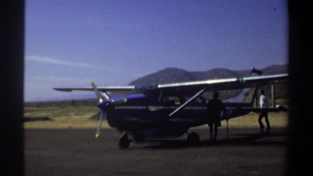 1969 Khartoum home-movie frame showing a blue and white single-engine plane on tarmac with people nearby and mountains behind.