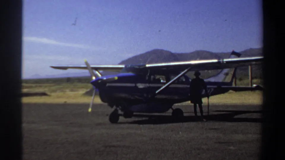 1969 Khartoum airfield frame showing a blue and white small plane, visible strut and propeller, with one figure beside the fuselage.