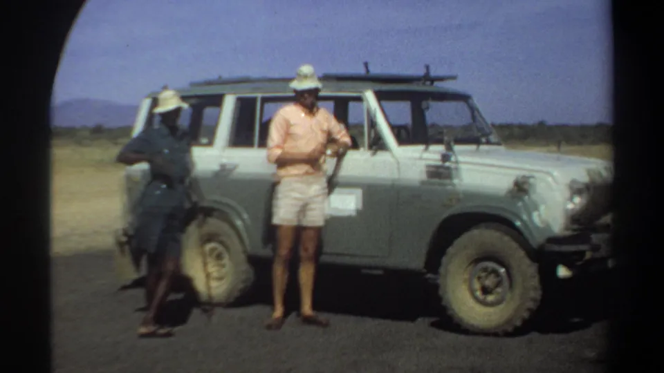 A 1969 Khartoum-area home-movie frame shows the civilian standing frontally beside the off-road vehicle while the uniformed man stays near the rear.