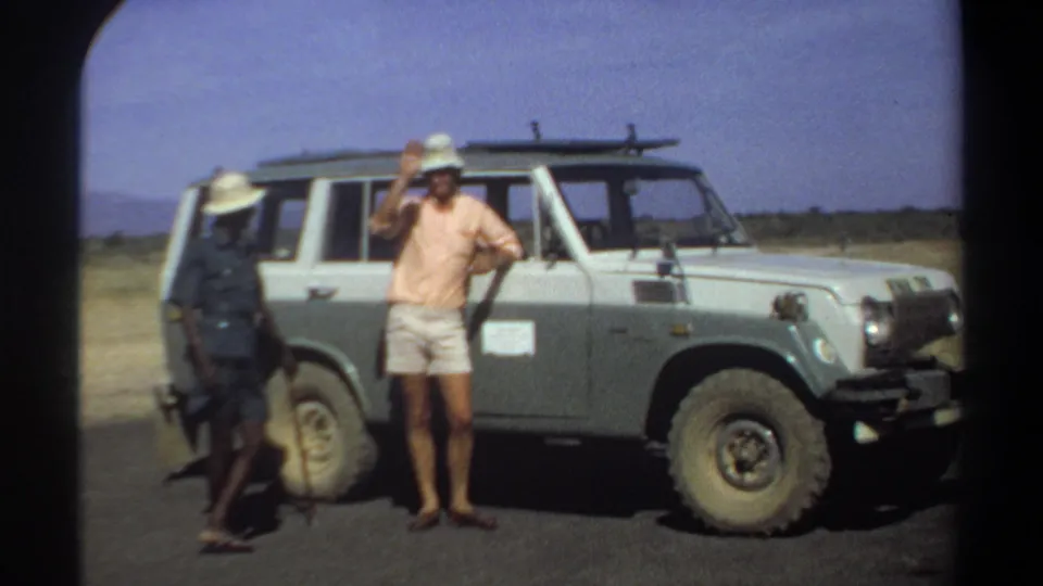 The civilian raises one hand near his hat beside the two-tone off-road vehicle, with the uniformed man still visible at the left.
