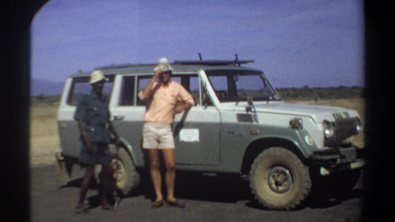 In 1969 Khartoum, Sudan, three men stand near a pale utility vehicle with a roof rack under a bright blue sky.