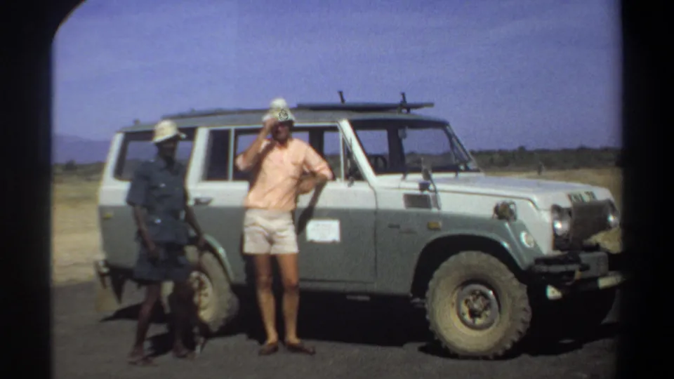 In 1969 Khartoum, Sudan, three men stand near a pale utility vehicle with a roof rack under a bright blue sky.