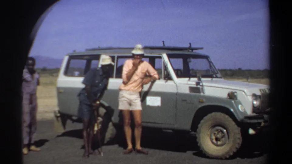 Three men remain beside the vehicle in the Khartoum home-movie footage, with one figure at the left edge of the frame.